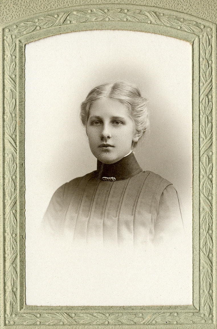 En okänd ung kvinna i mörk, höghalsad blus med stråveck. Vid kragen syns en brosch.
Bröstbild, halvprofil. Ateljéfoto.