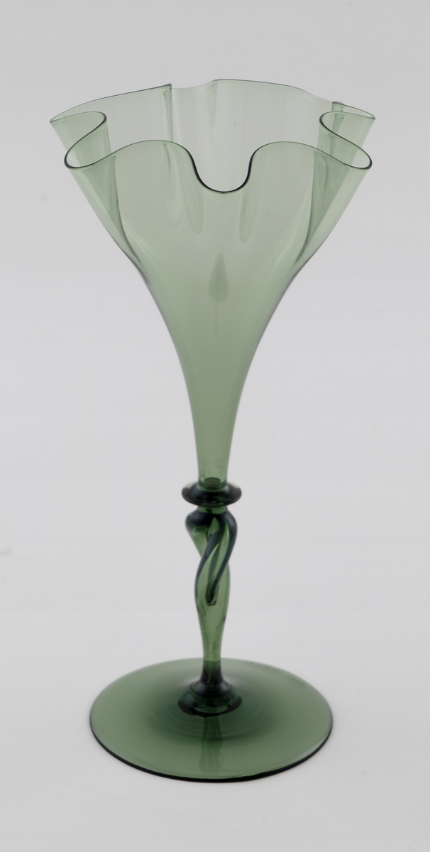 Glassvase i gjennomskinnelig, mørkegrønt glass. Konisk kupa med bølgeformet munning, som bæres av en spiralformet stett med vulst. Sirkulær fotplate med puntemerke, noe hevet i midten.