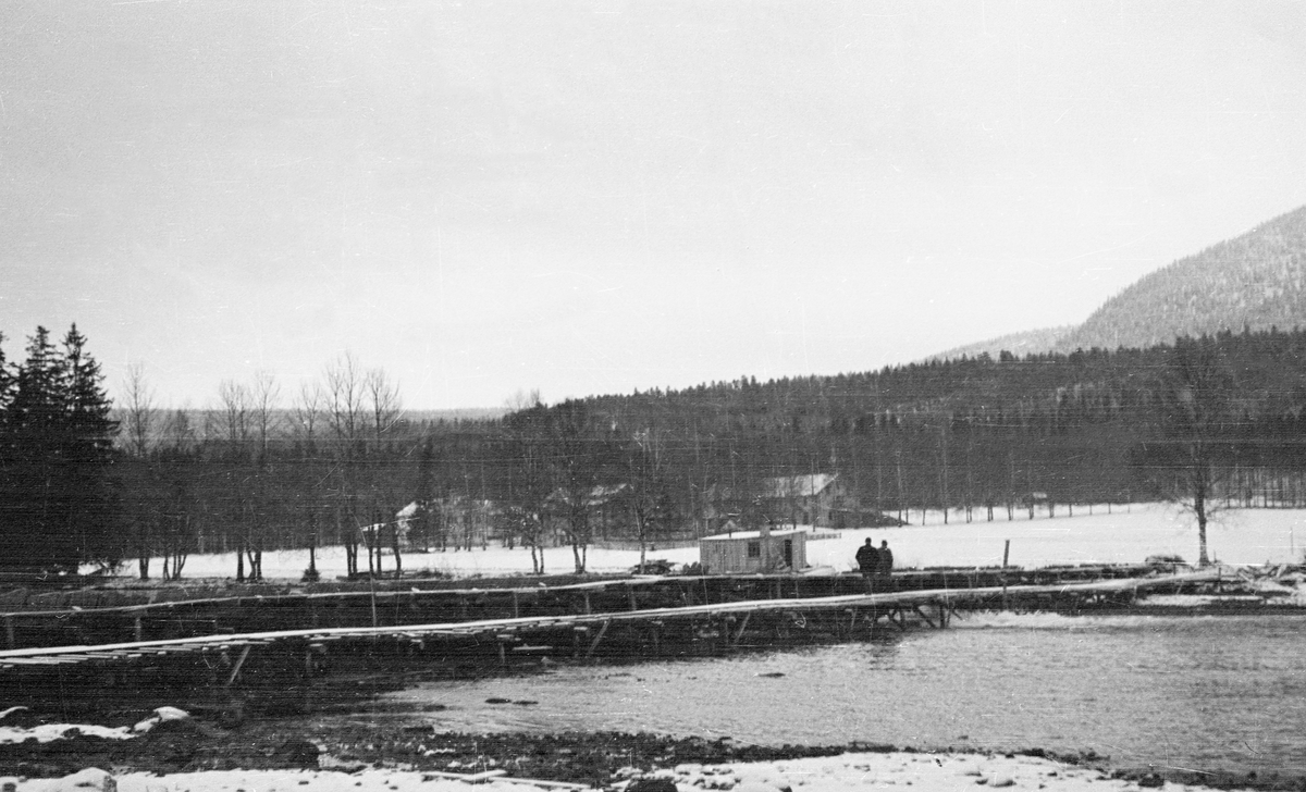 Ny damkonstruksjon ved Storsjøens utløp ved Løsset i Åmot, fotografert i desember 1941.  Fotografiet er tatt fra vestsida av vassdraget og det viser den nye dammen på skrå nedenfra.  Konstruksjonen består av en plankeskjerm som er montert på bukker på motstrøms side.  Plankeskjermen og bukkene støttes av en steinfylt trekonstruksjon.  Fotografiet kan tyde på at dette også er en slags bukke- eller stolpekonstruksjon.  To menn står på en bordgang oppå dammen.  Bak dem skimtes et lite hus, som antakelig tjente som arbeidsbrakke under arbeidet med dammen.  Bak dette igjen ligger tunet på garden Løsset på flate, snødekte jorder med barskogkledde høydedrag i bakgrunnen. 