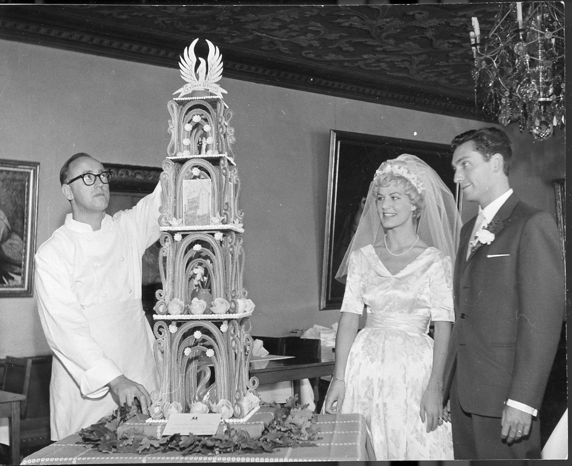 Ett brudpar tittar på en hög bröllopstårta. Konditorn, Knudsen, står intill densamma. Överst en logo med text: "Gyllene Uttern". De befinner sig i Gyllene Utterns nedre matsal.