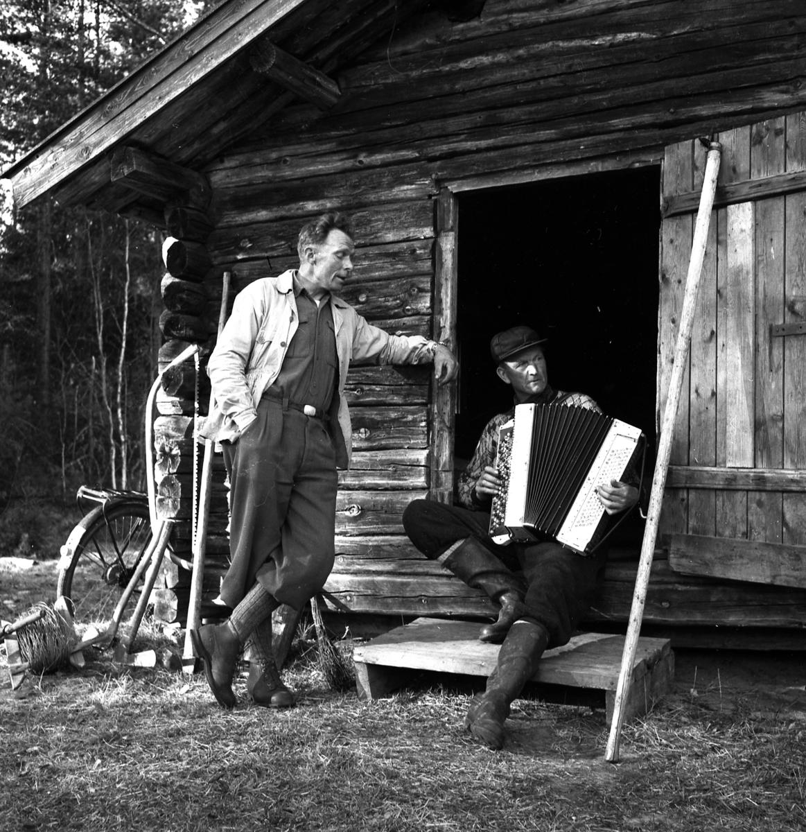 ​To menn ved et lafta hus. Den ene står lent mot dørkarmen, den andrre, Johan Elsmo, spiller trekkspill. Bildet er tatt ved fløterdammen på Jømna.
