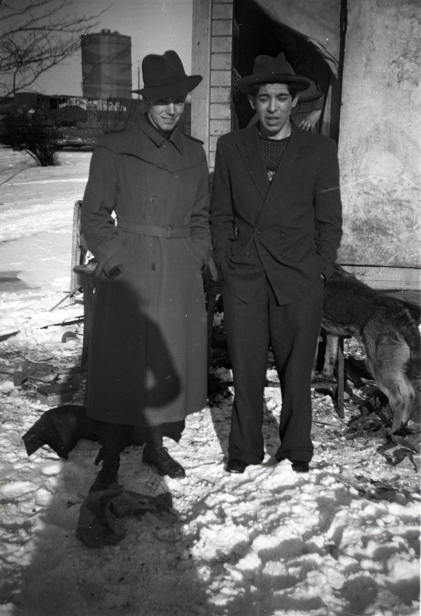 Två män poserar för kameran stående i snön. I bakgrunden syns Göteborgs gasklocka. Bilden är troligen tagen vid den romska lägerplats som fanns i Bergslagsparken i Göteborg på femtiotalet.