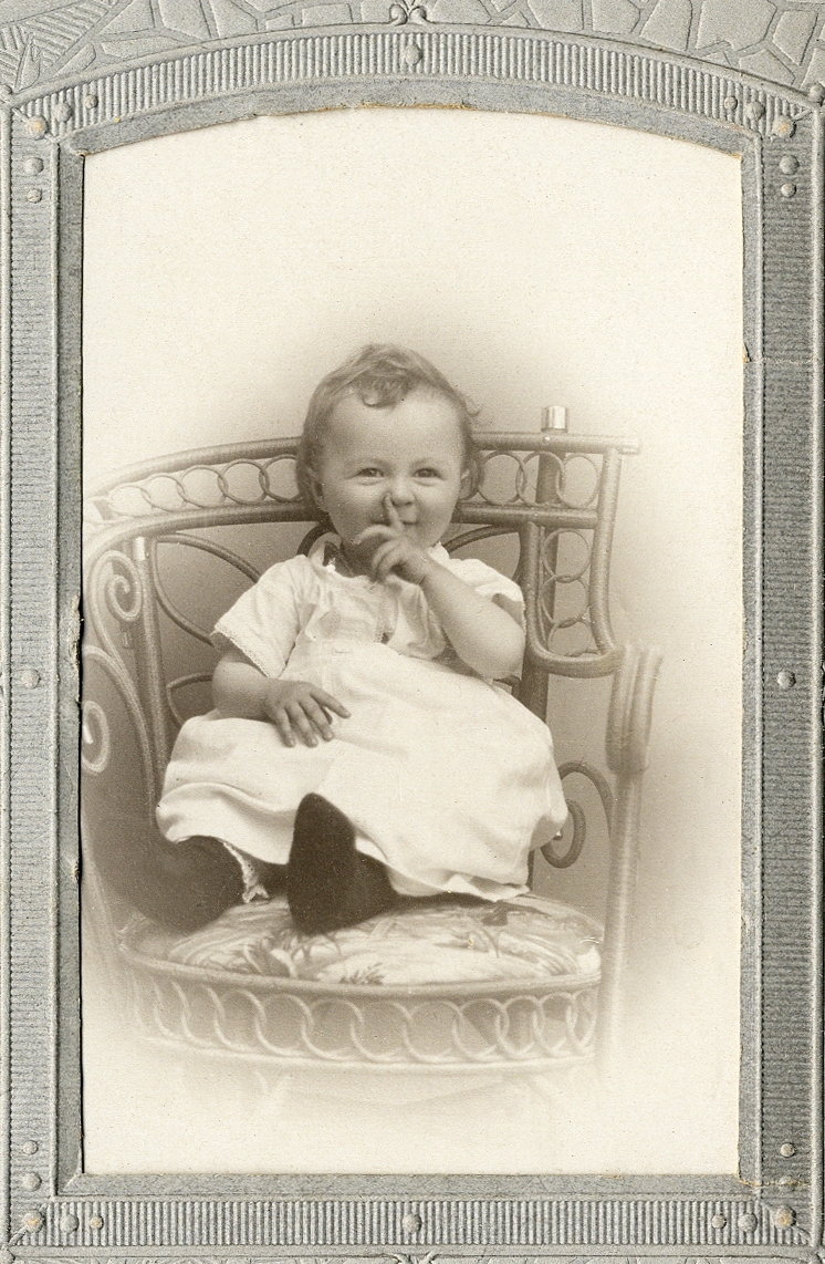 En liten glad flicka i klänning/kolt som sitter i en rottingstol. 
Helfigur. Ateljéfoto.

Fotografens dotter.