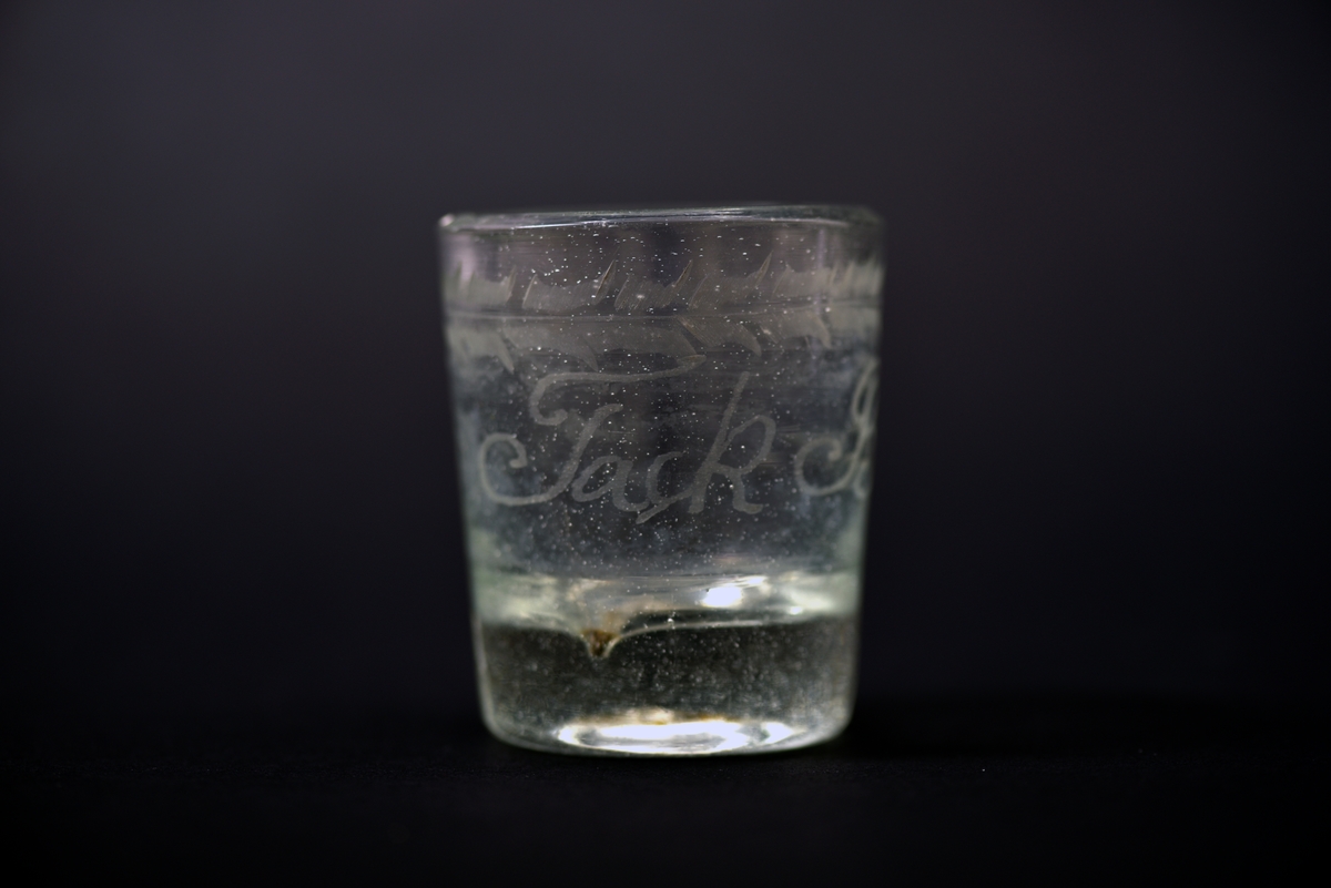 Dricksglas i något konad form av ofärgat, tjockt, glas. 
Dekorerad med en graverad bladbård vid mynningen och därunder en inskrift "Tack Bror."