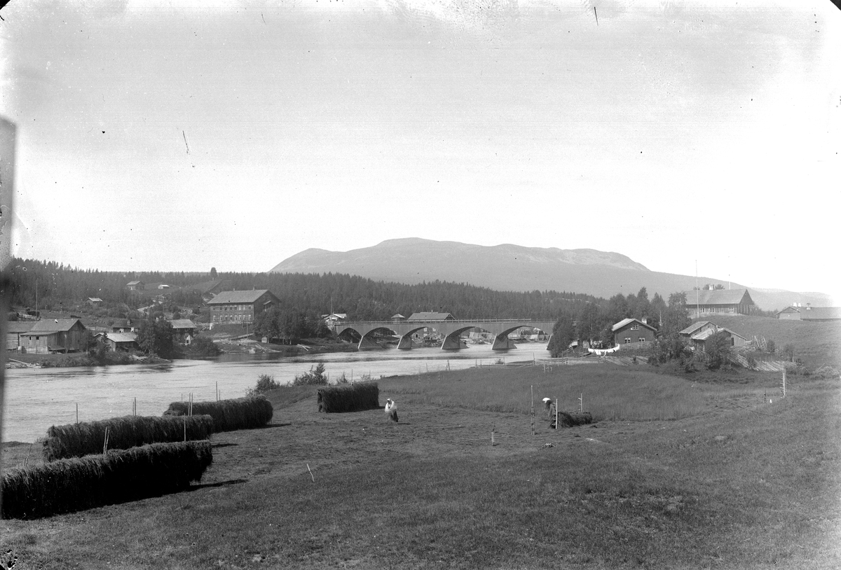 Nybergsund. Fotografert fra østsida av Trysilelva mot nordvest, Trysilfjellet i bakgrunnen. Brua sto fra 1879-1929. På vestsida av elva ses Nybakk og Nyberg, huset i to etasjer på østsida som ligger delvis skjult bak en høyde er Folkvang.