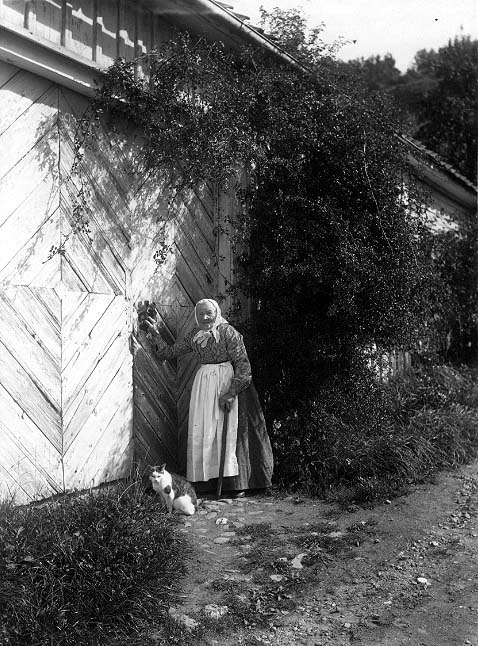 Hushållerskan Augusta Jakobsdotter och katten Tasse står i gränden vid porten till Franckska gården. Augusta bär ljust huckle och förkläde och håller en käpp i vänster hand. Det är sommar med rik växtlighet utmed husfasaden.