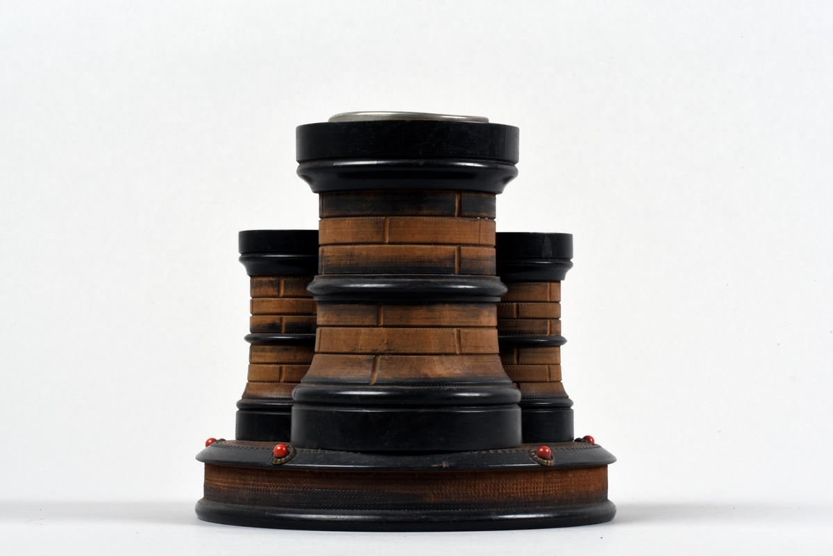 Cigarrställ av trä, i form av tre runda torn som står på en rund platta. Det högsta tornet har en metallskoning vid mynningen, de lägre olika utformade hål.