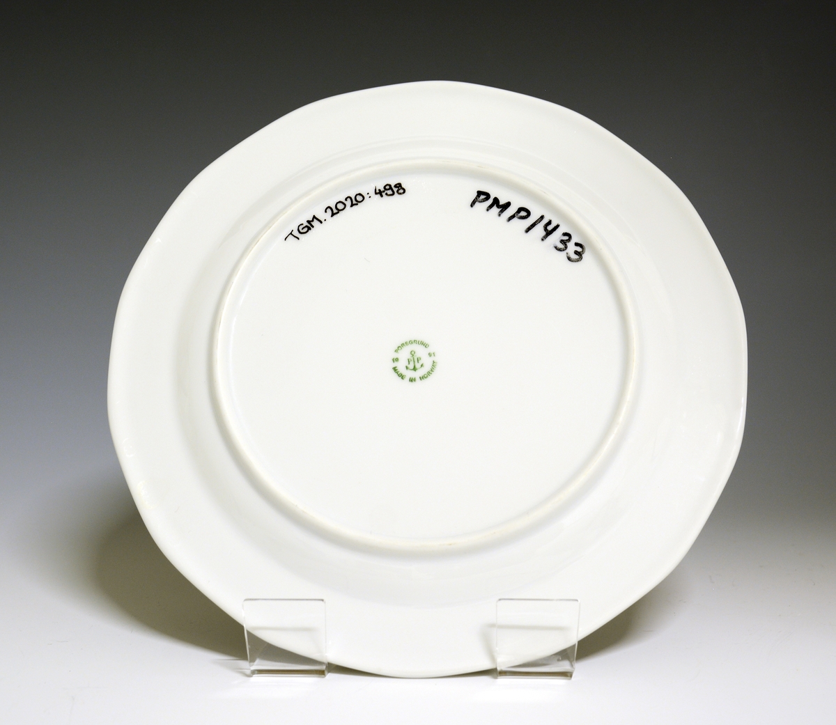 Mangekantet tallerken av porselen med hvit glasur. Dekorert med en bord ytterst på fanen med blomster og geometriske former. 
Modell: Octavia, i produksjon fra 1977
Dekor: Innkjøpt dekor, ikke av Grete Rønning.