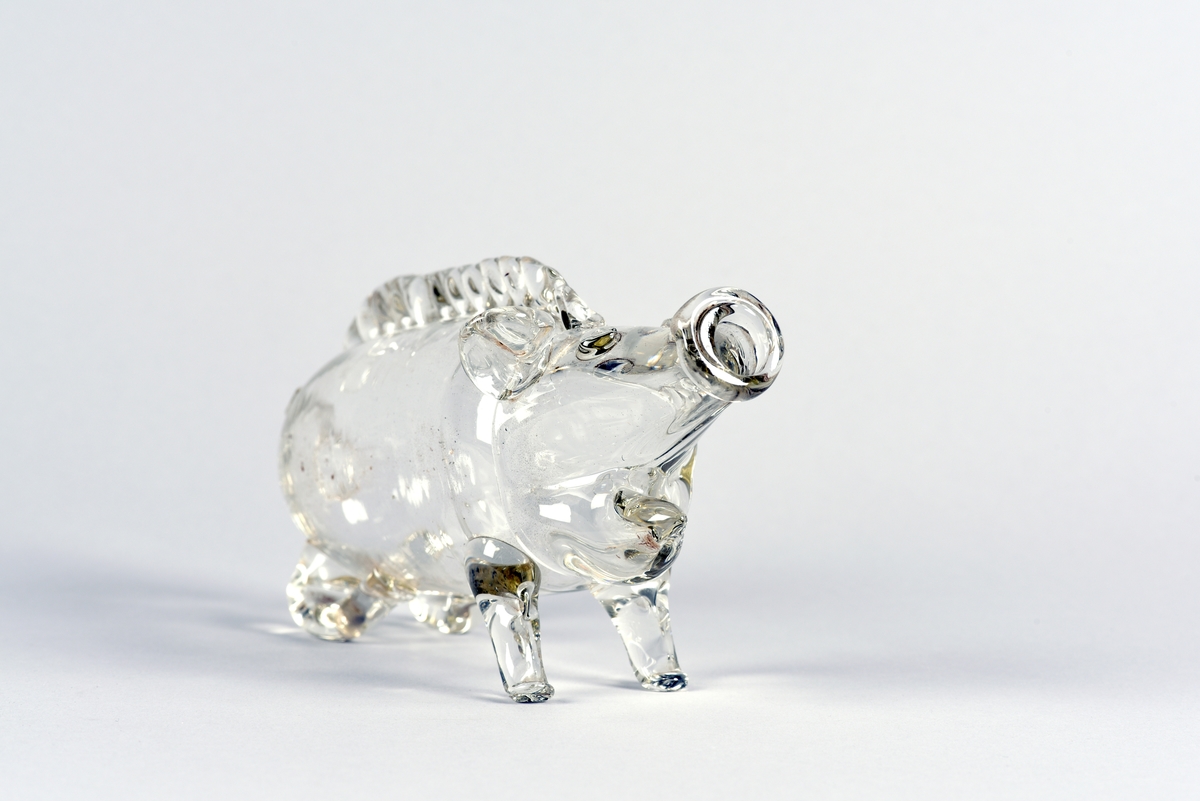 Brännvinsflaska i form av en gris, så kallad fyllesvin. Tillverkad av klarglas. Halvsittande gris, där trynet fungerar som flaskmynning. På ryggen upprättstående ragg.