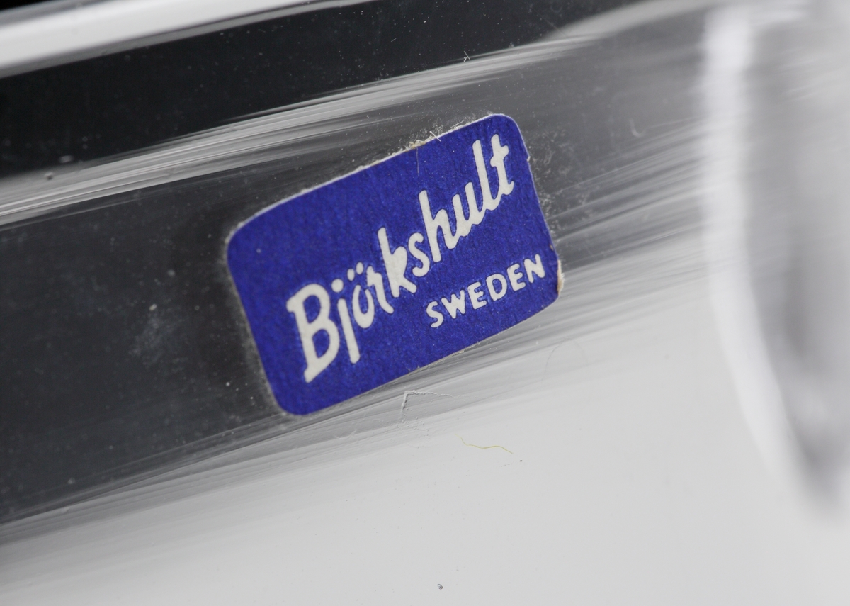 Satt modell bestående av en rund nedre del som övergår i en cylindrisk hals/mynning med isklippt pip. 
Etikett: Blå botten och vit text "Björkshult Sweden".