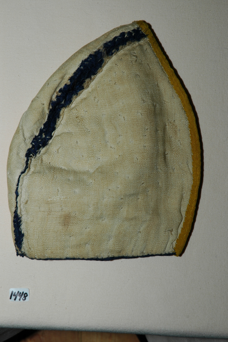 Lue, tobladlue av mørkeblå silkedamask kantet med knallgult silkeband (lerretsvevd). Lua er fôra med linlerret og vattert med stry. Nedrekanten er sydd til med faldesting med grått lingarn. De to delene er sydd sammen med attersting. Sømmonnet er lagt til side og kastet sammen. Kantebandet er vrangsydd på med attersting på retta og faldesting på vranga, 0,5-0,8 cm bredt. Langs sammensyingssømmen er det satt på to rader med en ca. 2 cm brei knipling. Registrering ved Bunad- og folkedraktrådet/Norsk institutt for bunad og folkedrakt v/Jon Fredrik Skauge, 2005, revidert v/Bjørn Sverre Hol Haugen, 2020.