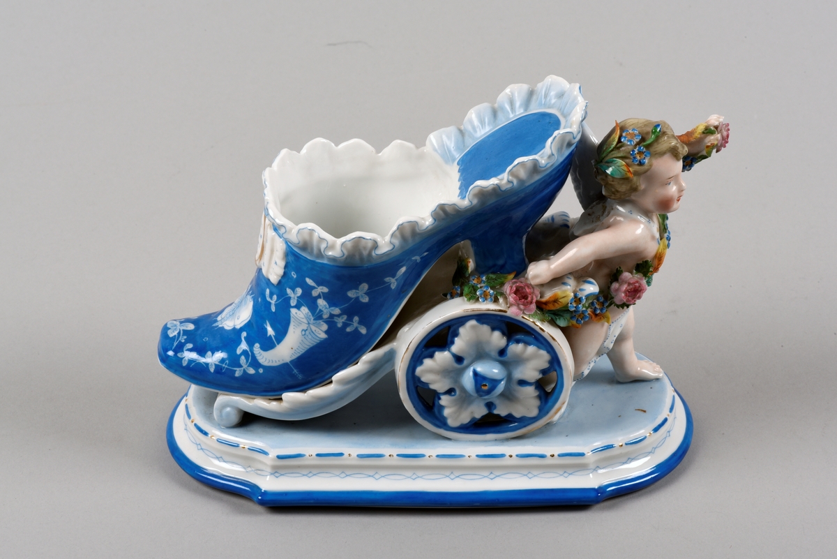 Prydnadsföremål, vas, tillverkat av bemålat vitt porslin. Vasen är i form av en sko på hjul, som dras av en putti. Selen består av en blomstergirland.