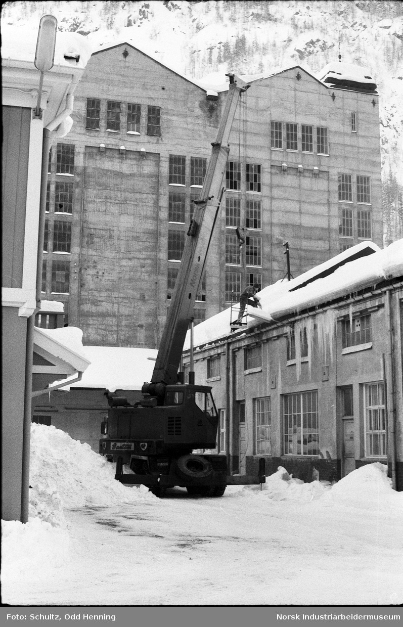 Folk som rydder snø på takene på fabrikkbygning med hjelp av en lift.