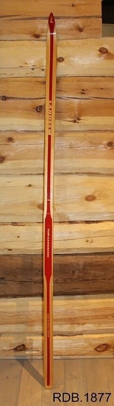 Avsmalende jevnbred ski med liten bue. Bøy uten tupp. Avfaset bakende. 
Dekorert med rød stripe på trehvit bunn.