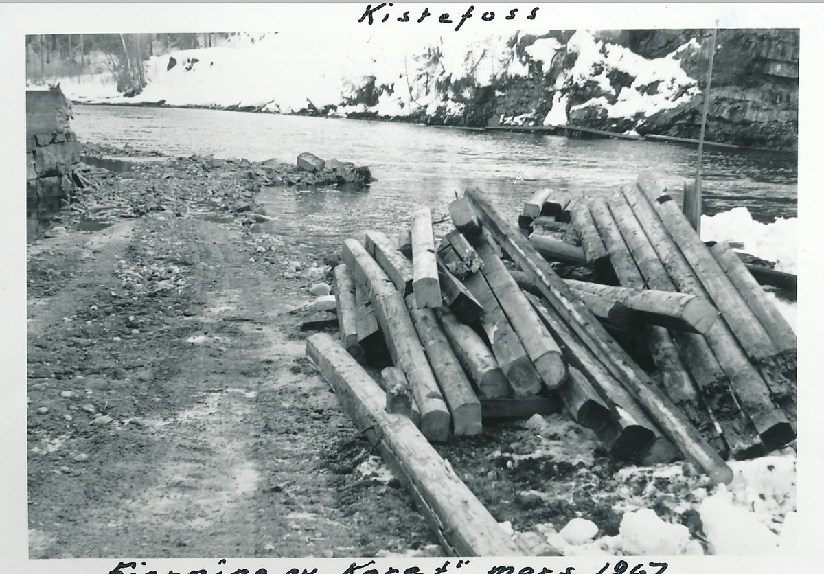 Tilhugde tømmerstokker ligger i stabel på elvebredden ved Kistefos. Anleggsvei til venstre i fotografi.