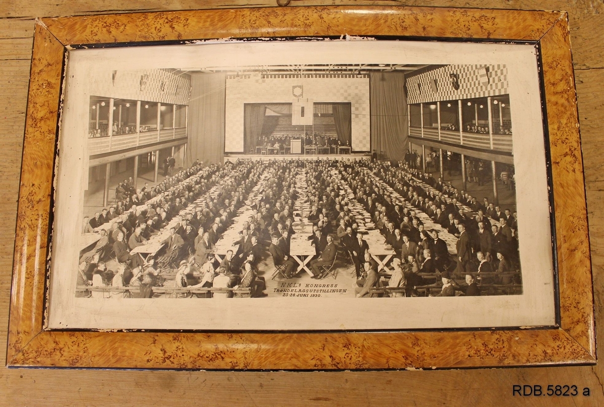 En stor forsamling menn og noen kvinner, deltakere på NKL's kongress under Trøndelagsutstillingen i 25.-28. juni 1930. Mennene sitter med papirer foran seg langs 7 lange bord . Det er også en scene fremst i salen og balkonger på hver side hvor det også sitter mennesker.