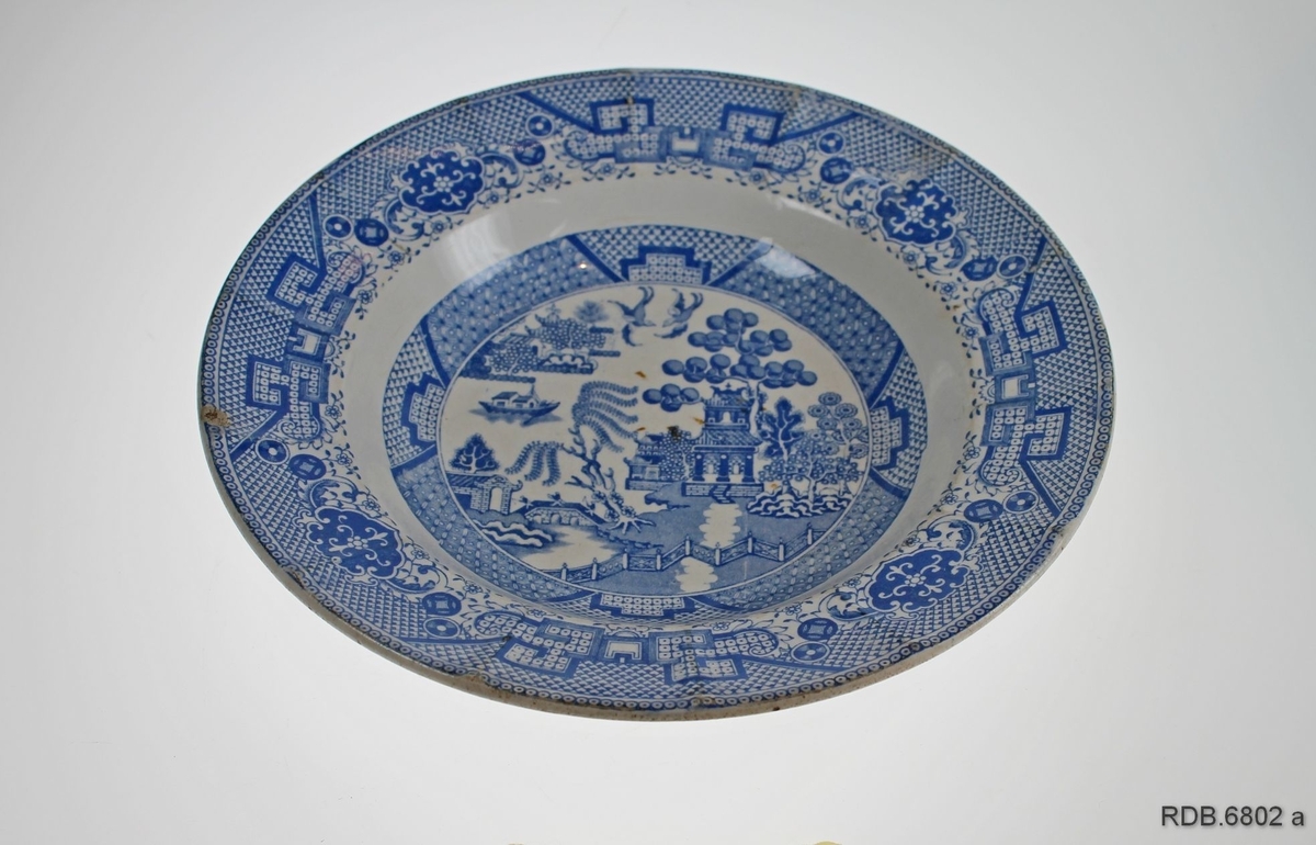 Flat tallerken med mønsteret "Blue Willow", også kalt "China" i blått på kvit bunn. Et skår i kanten. Fabrikkstempel under.