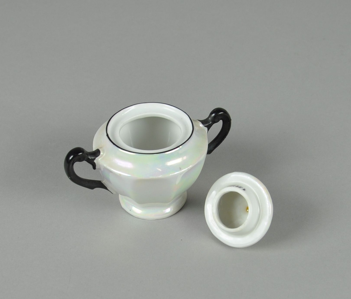 Sukkerkopp av glassert keramikk, med lokk. Koppen har to hanker. Koppen har perlemorsflate, med svartfarget maling på hankene, ved munningsrand og knott på lokket. Koppen har fassetert profil.