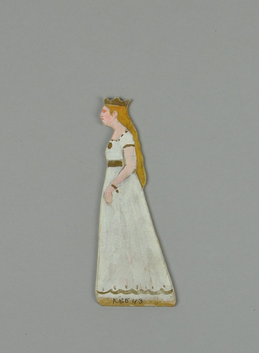 Figur av papp. Figuren forestiller stående dame i sideprofil, med krone og hvit kjole. Figuren er malt på begge sider.