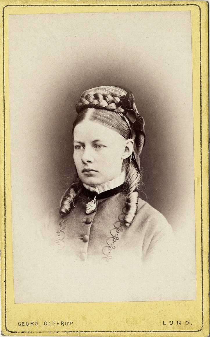 Foto av en kvinna i ljus klänning med knappar och sammetsgarnering och ljus kråskrage. Vid kragen syns en brosch. 
Bröstbild, halvprofil. Ateljéfoto.