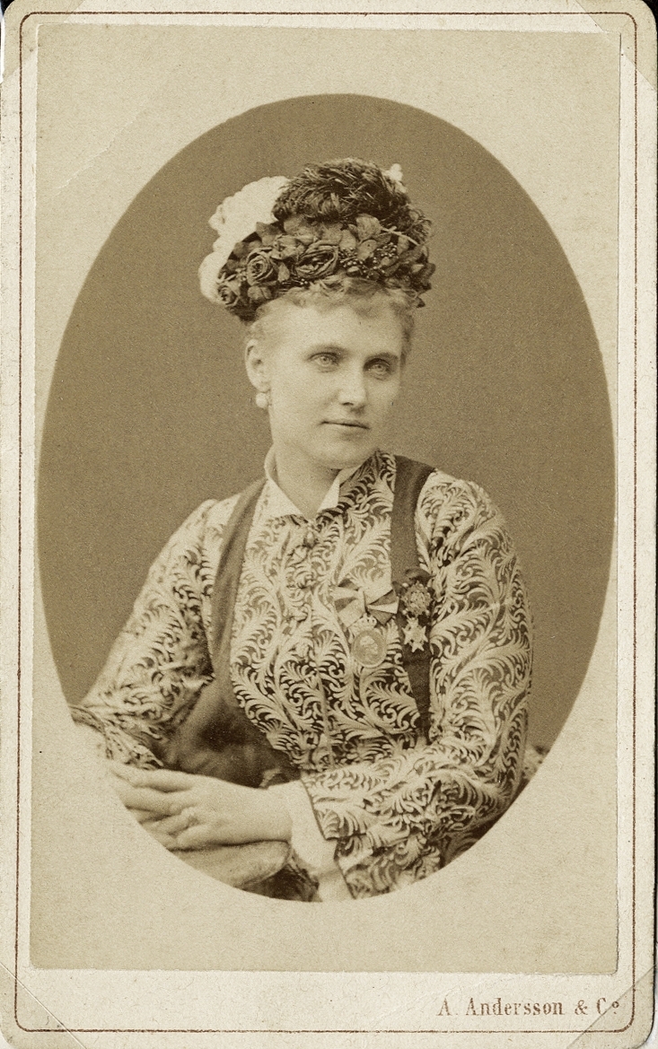 Foto av en kvinna i mönstrad klänning med en ornerad hatt på huvudet, prydd med fjädrar m.m.
På bröstet skymtar några ordnar och medaljer. 
Midjebild, halvprofil. Ateljéfoto.