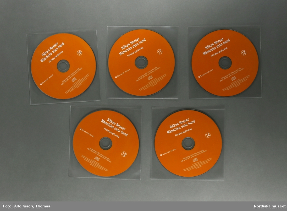 CD-bok.  a:1-2-n:1-2) 14 st CD-skivor, i en kvadratisk, transparent plastficka, innehållandes uppläsning av romanen "Människa utan hund" skriven av författaren Håkan Nesser som även är uppläsaren på denna CD-inspelning.  Inspelningen är totalt 15 timmar lång.

CD-skivorna är tunna cirkelfornade,  12 cm i diameter, på ena sidan oranga med text om titel samt kort om produktionen. Varje skiva är numrerad för att visa rangordningen i vilken skivorna ska lyssnas på/spelas. Baksidan på skivan är silverfärgad, speglliknande, och innehåller själva ljudspåren som läses av när skivan placeras i en CD-spelare.
Varje skiva är separat plackad i en kvadratisk, transparent, plastficka för att skydda mot repor som ev skulle kunna skada skivan. 
Samtliga skivor är tillsammans packade i en o) kvadratisk papperskartong/ konvolut. Konvolutet har på framsidan en bild av ett fönster med ett gultonat fotografi visandes ett fönster med en flaxande fågel utanför. På konvolutets baksida finns en introduktion till bokens handling samt  information om tillverkningen av denna CD-bok.
(CD är en förkortning av engelskans Compact Disc. CD-skivan kan lagra digital information, exempelvis musik.
CD skivans konstruktion: överst är ett transparant plastlager följt av ett tunt lager av blank metall med ett präglat spiralformat spår med mikroskopiskt små gropar som avläses av CD-spelarens laserstråle när skivan snurrar (ca 500-700 varv/minuten). Ljuset studsar tillbaka och läses som en följd av ettor och nollor, som i detta fall, blir till ljud.(Källa/. Nationalencyklopedin)


/Cecilia Wallquist 2019-02-20