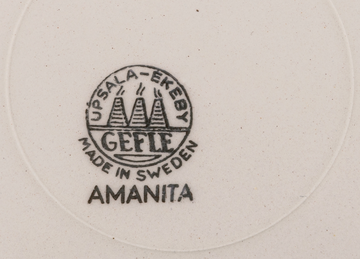 Assiett (med tillhörande kaffekopp och fat) tillverkad vid Gefle Porslinsfabrik AB. Röd bottenfärg med vita prickar. Dekorens namn "Amanita" formgiven av Helmer Ringström. Koppens modell förmodligen formgiven av Arthur Percy.
