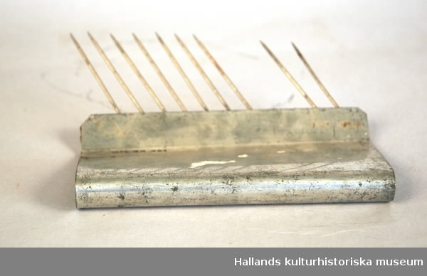 Kakstöd av metall, som används som stöd vid perforering av sega kakor medcellperforator. Hopvikt metallskiva som avslutas i ett veck i vilket vassa metallnålar är fastsatta. Saknas: tre nålar.