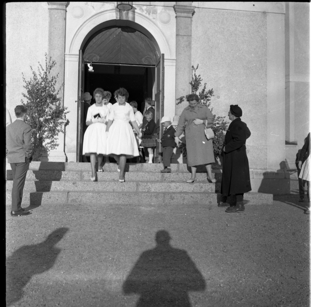 Kvinnliga konfirmander på väg ut ur Gränna kyrka. Vid trappan står barn och vuxna. I förgrunden fotografens skugga.