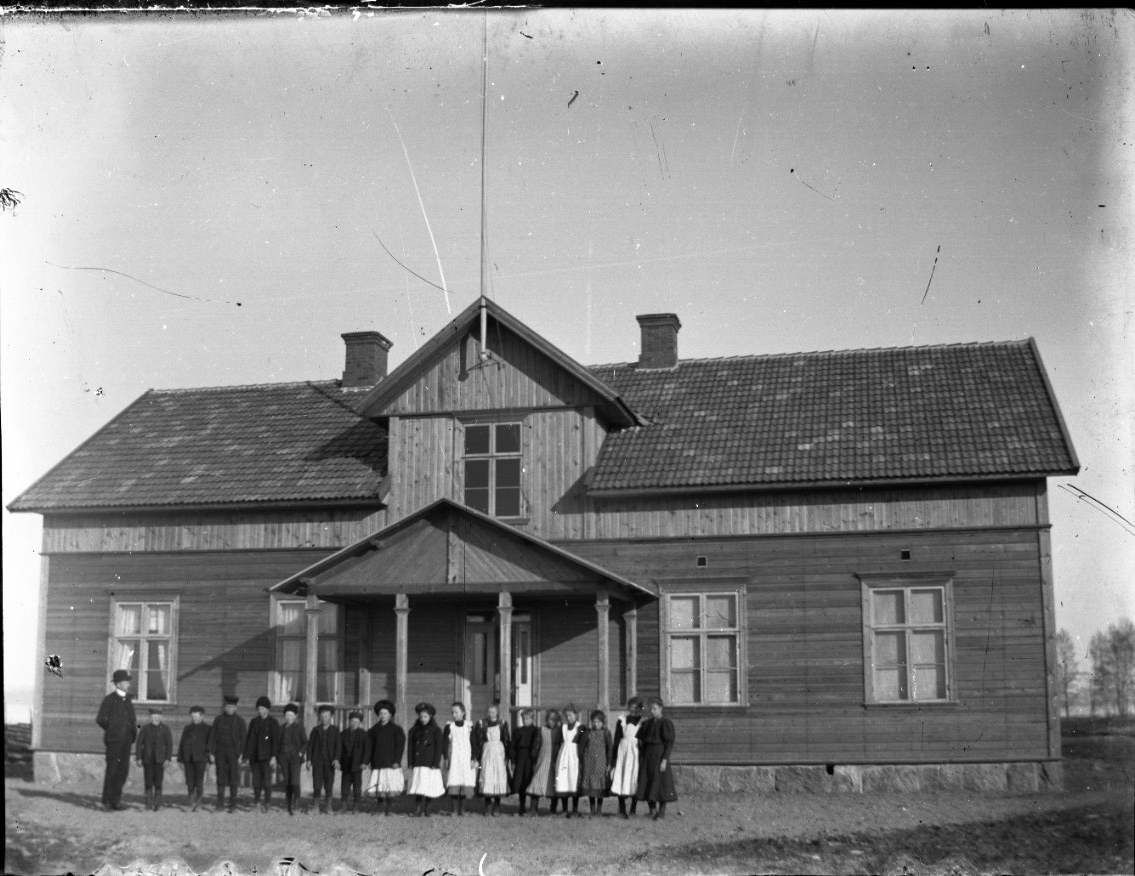Barn på rad framför en större träbyggnad, flickorna har förkläde. Sannolikt en skolklass. Byggnaden har liggande panel på bottenvåning samt stående under takfot och på frontespis.