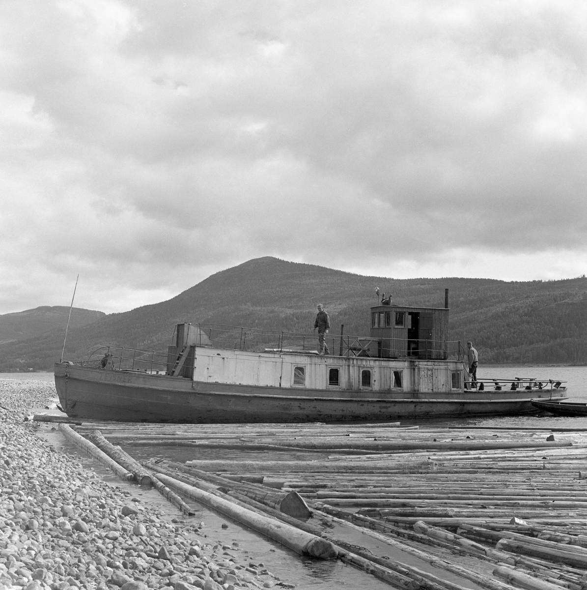 Slepebåt ved innsjø. Tømmersleping med slepebåten M/S Teisner på Fyresvatnet, Fyresdal, Telemark. Skipper Gunnar Sandvik . Båten ble bygd på Akers mekaniske verksted, og var opprinnelig en kombinert passasjer- og slepebåt fra 1876, Den fikk navnet M/S Teisner i 1913. Etterhvert ble det en ren slepebåt. Etter at tømmertransporten forsvant i 1971, ble båten tatt vare på av "Reiarlaget Teisners vener", men i 1990 ble båten dratt på land. Fyresvatn.