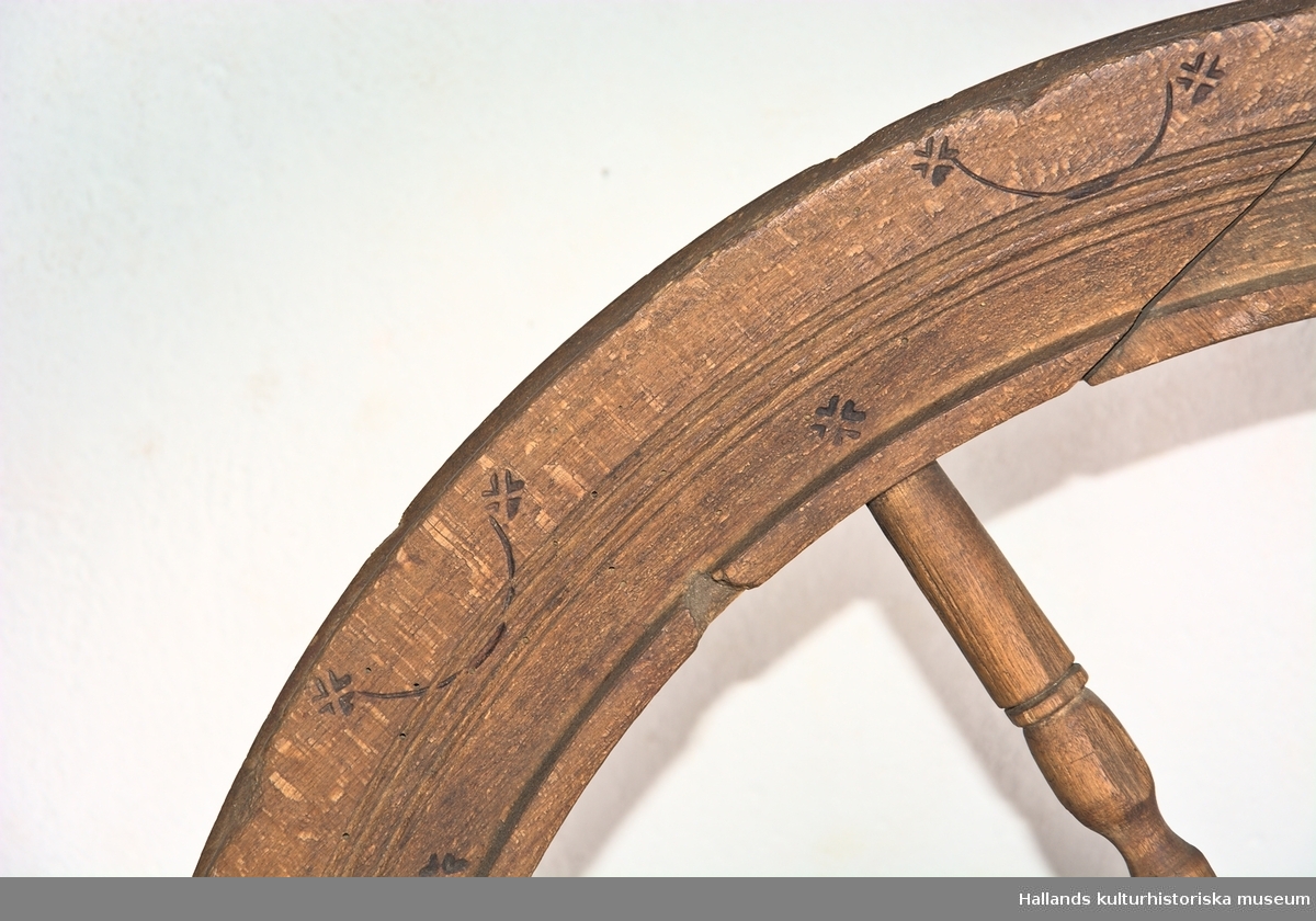Spinnrock av trä med axel av järn och lager för rullen av läder. Snedställt bröst. Hjulet vilar på dubbla stöd. Inbrända stjärnmönster och bågar på bröst och hjul. Mått: hjulets diameter 29,3 cm, hjullötens djup 6,4 cm. 