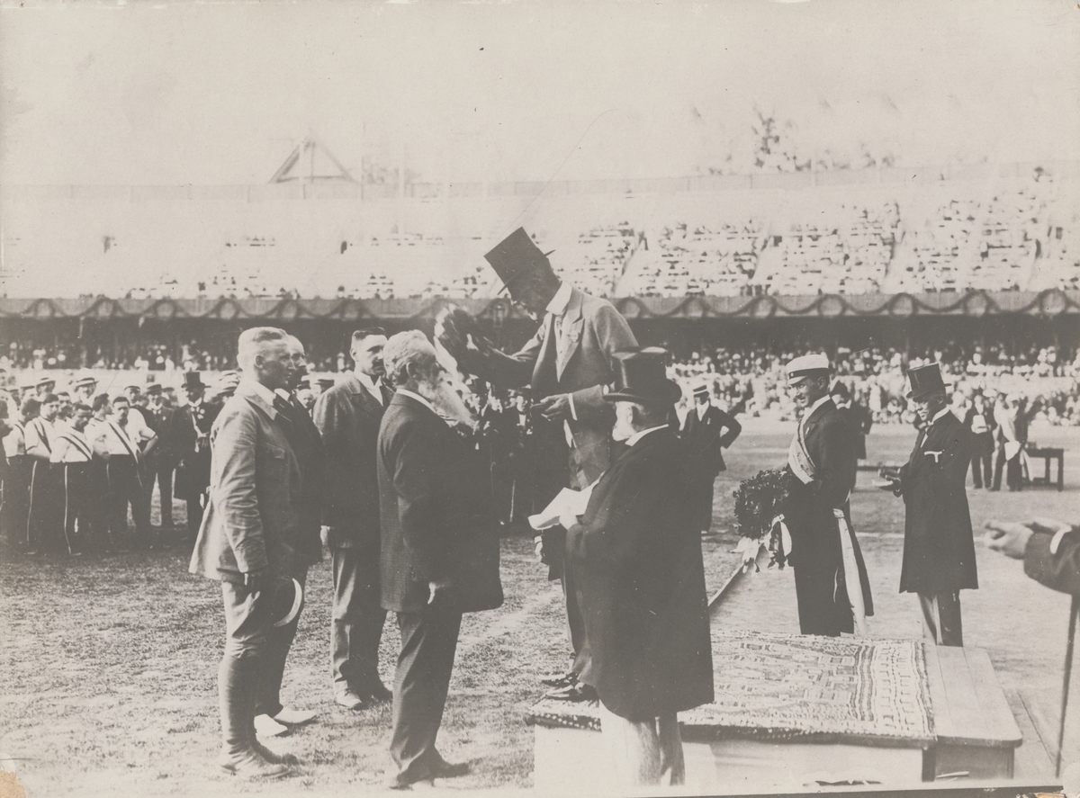 Grupp M V.

Skytten Oscar Gomer Swan tilldelas medalj av kung Gustav V under Olympiska spelen i Stockholm 1912 för segern i löpande hjort enkelskott, lag. Guldmedaljen från 1912 som Oscar Swahn vann vid en ålder av 64 år och 257 dagar gör honom till tidernas äldste olympiske guldmedaljör.