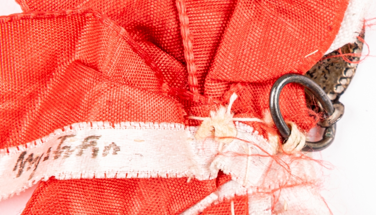 Ovalt hänge i rött och vitt sidenband, sammansydda med handstygn. Bandet märkt "Nyström".
Hänget stansat: "d. 17 Mars".