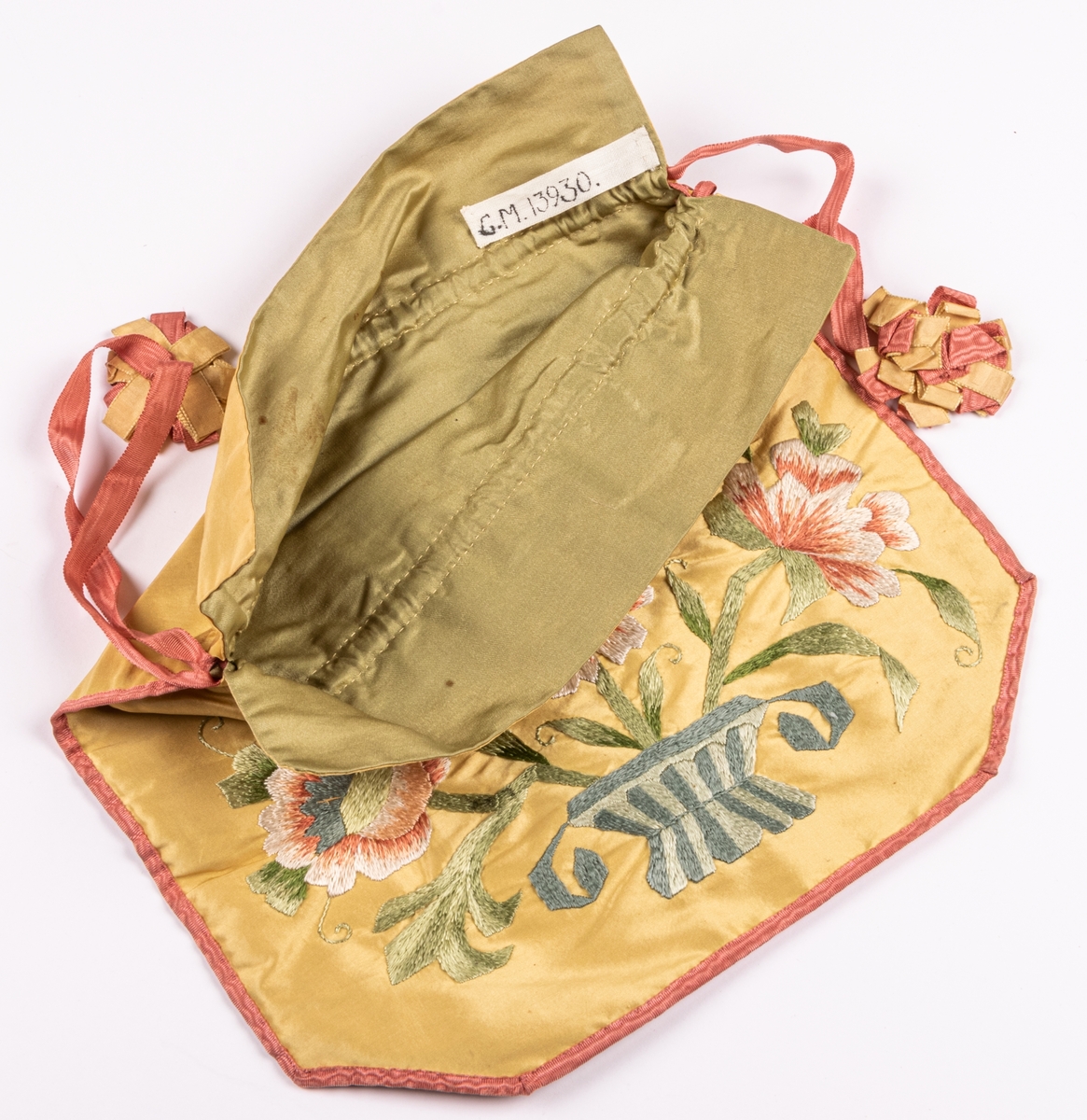 Väska av gult siden, broderad i schattersöm kantad med rosa ripsband som även utgör dragband och prydda med kokarder i rosa och gult, bodrad med grönt siden. Dragsko. 1890-tal.