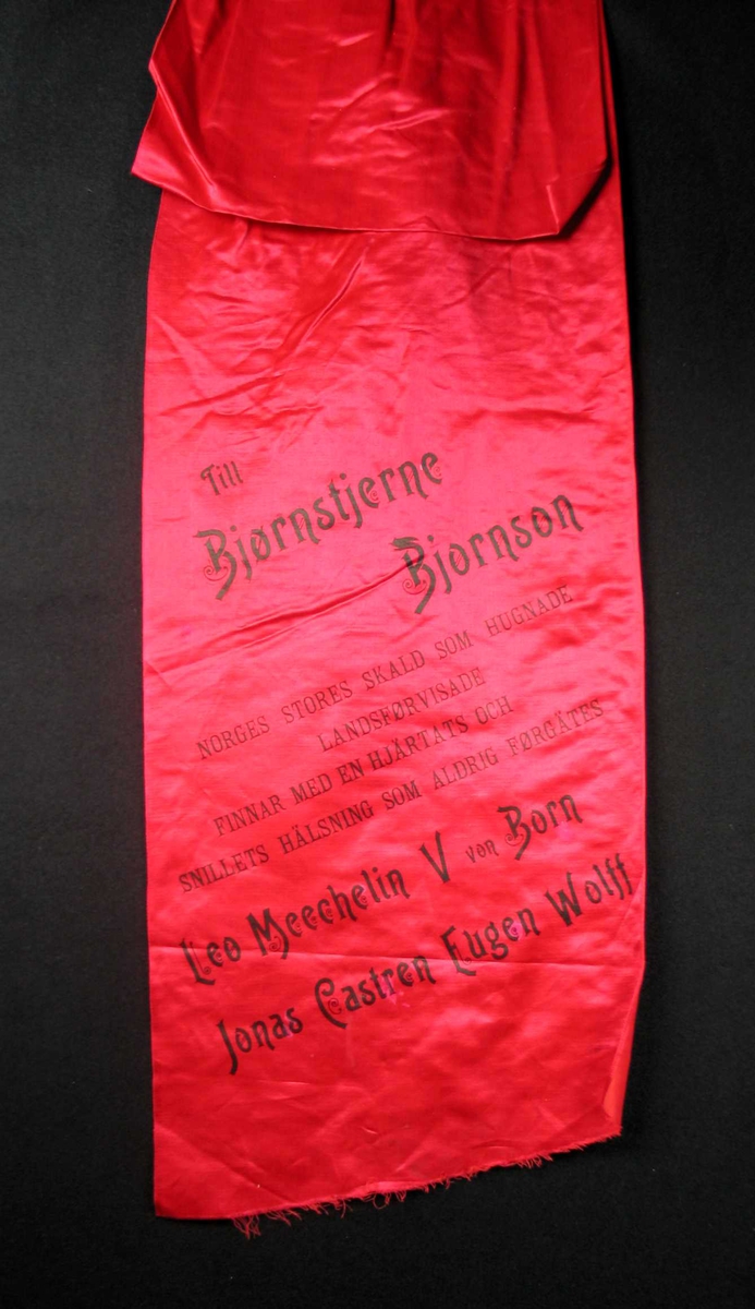 Begravelsessløyfe i rød silke med svart tekst.