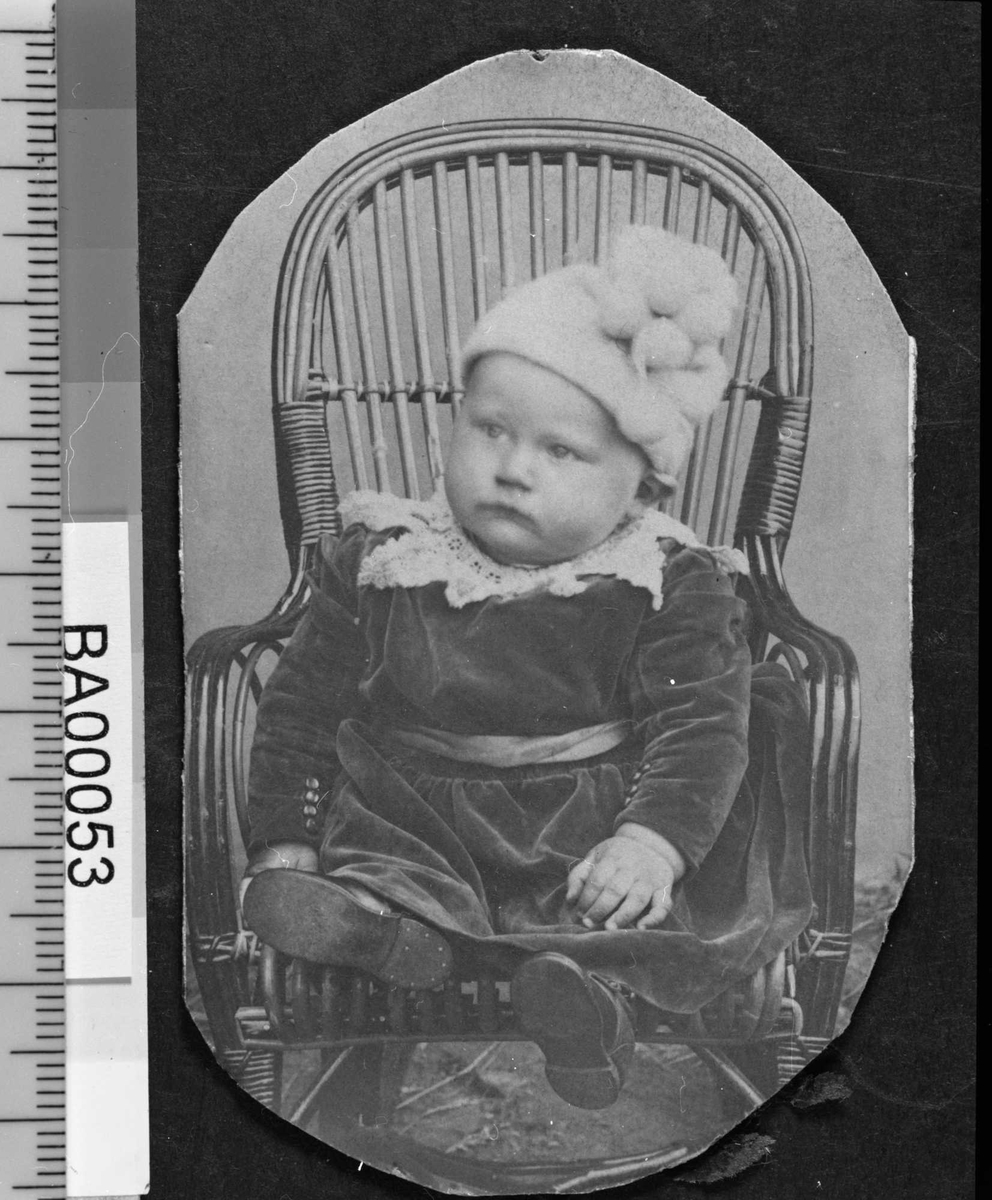 Fotografi av lite barn sittende i kurvstol med lys strikkelue med ponponer festet på siden, mørk kjole med stor hvitt kniplingskrave rundt halsen og sjerf rundt magen, små pynteknapper langs innsiden av mansjettermene. Barnet ser til høyre.