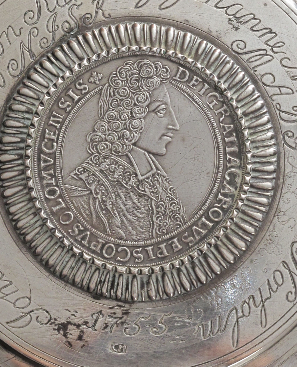 Stentøykrus m. lokk i sølv, lokket laget 1755.  
Flatt lokk m. buet, profilert kant, innsatt mynt  m. radiært riflet innr. grav. :   Johannes Salwesøn Kallevig,   Johanne NielsDatter Norholm, 1755,  samt   initialer for seinere eiere.  Sølvsmedstempel. 

Mynten profilportrett m. innskr: DEI GRATIA CAROLVS   EPISCOPVS OLOMVCE N(?) SIS"  reversen kronet våpen m.  ørner etc. :  DUX LOTHAR ET BAR S R I PCPS   RE CA BOKO 1704. 

Lokkgrep som stående blad m. opprullede fliker.  

Lokket montert på sylindrisk stentøykrus,  nederst brunt,  riflet feldt, derpå glatt, så lyst, riflet  feldt, buet,  profilert hank forts.