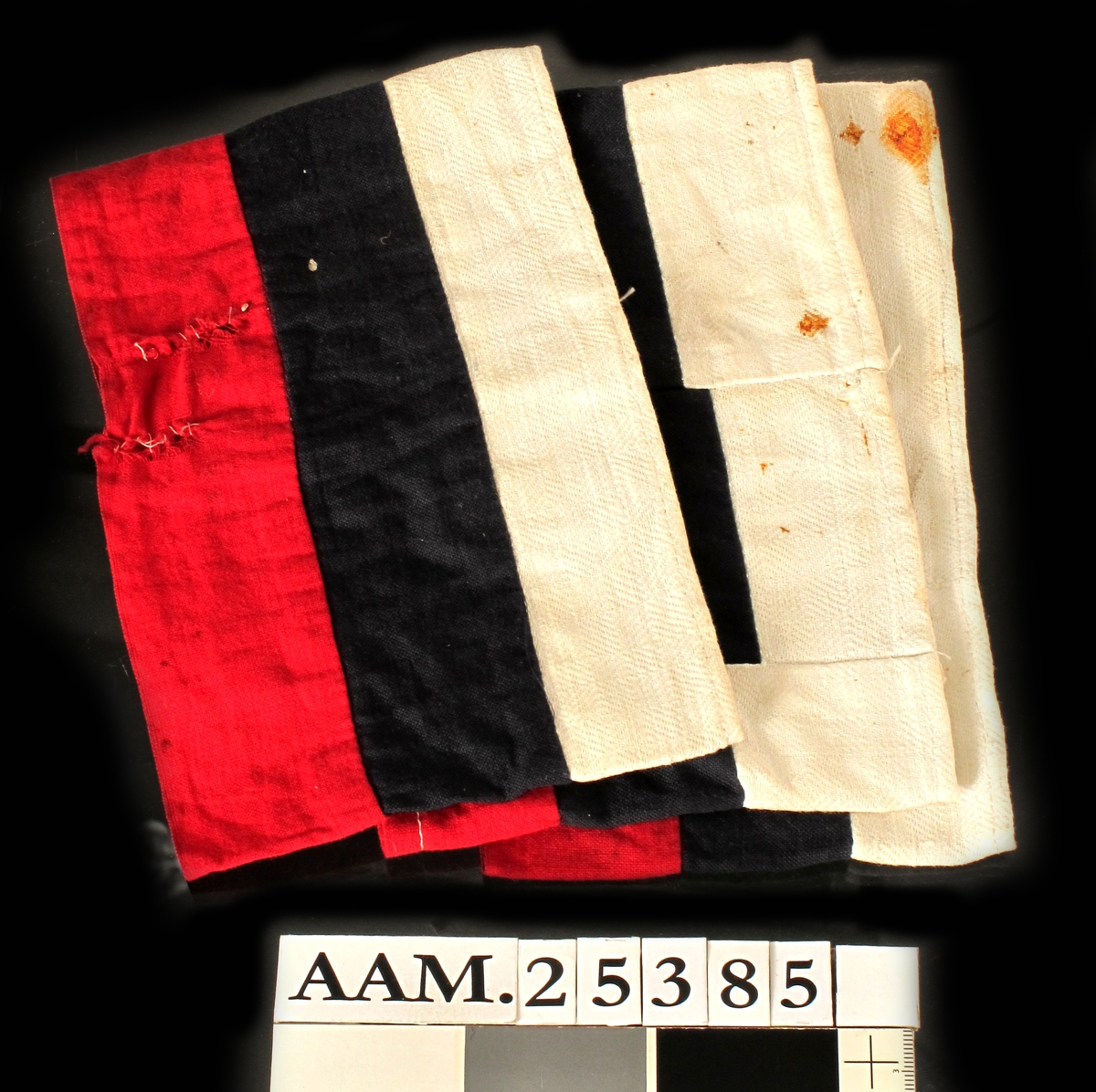 Armbind av bomullstøy, m. 3 horisontale striper i flaggets farger. Var ved innkomst utstyrt med en svært rusten sikkerhetsnål som ble fjernet.