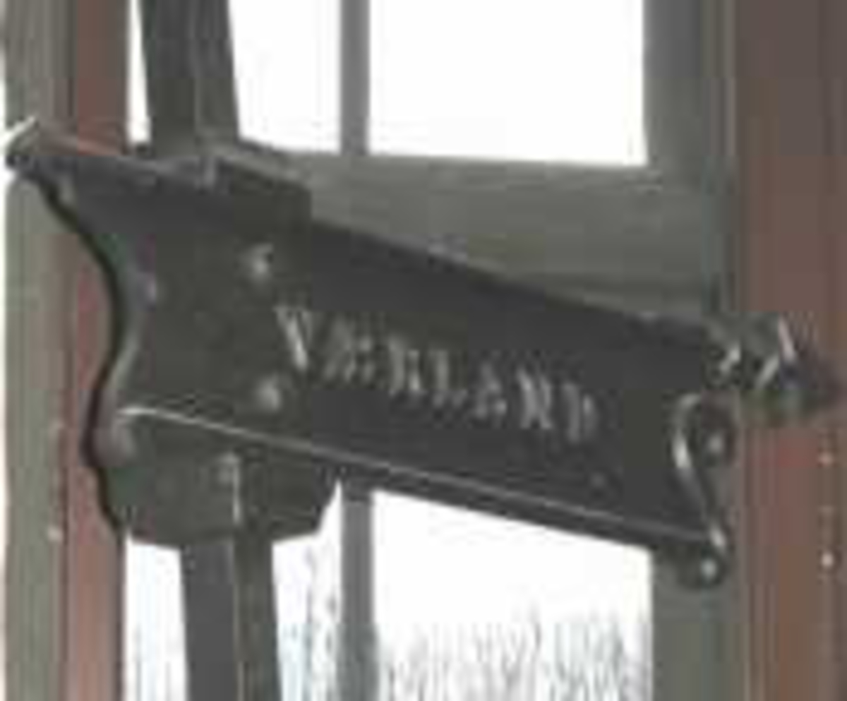 Stolpe  m  henvisningsskilt med tekst Værland. Selve skiltet er elegant utformet med enkel akantuspynt i begge ender. Skiltet er skrudd fast i stolpen. Kule på toppen av stolpen mangler.
