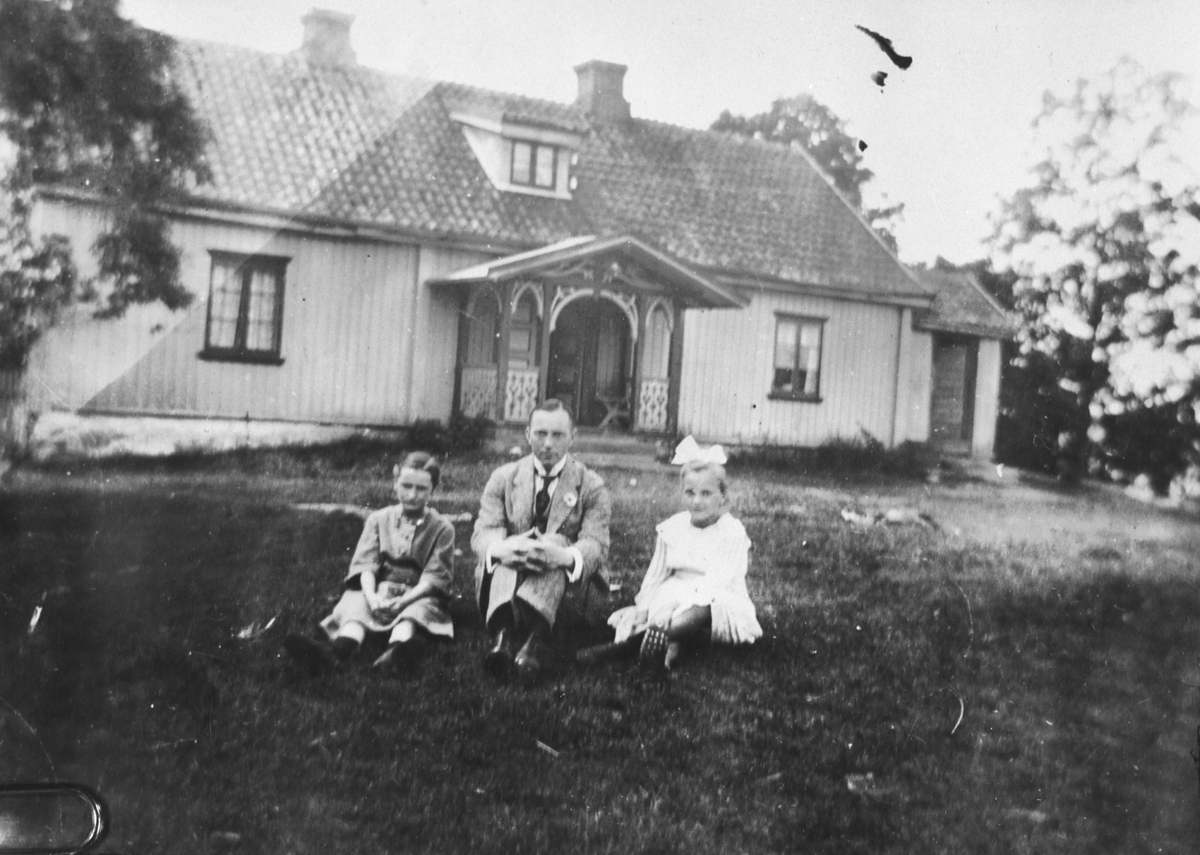 En mann og to jenter sitter i gresset foran et bolighus.