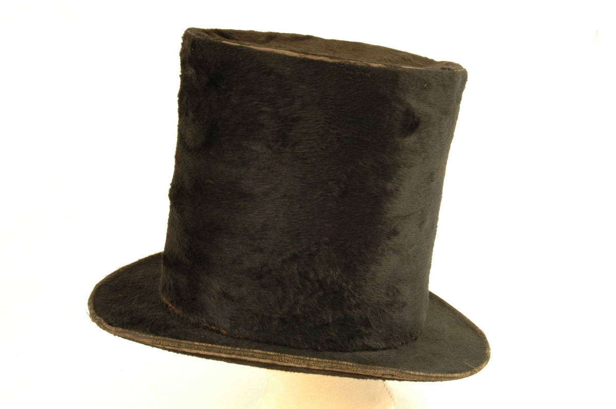 Svart flosshatt med høy pull og brem. Innvendig er hatten kledd med bred skinnbestetning.