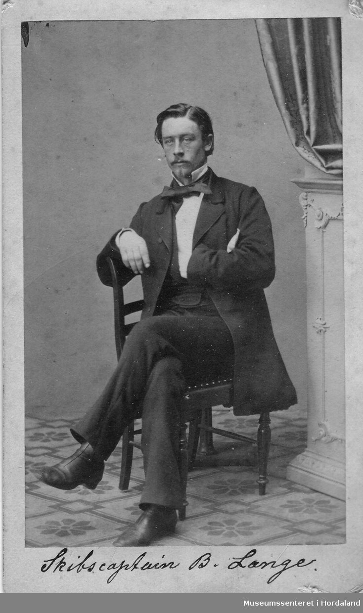 Mann med lang frakk, dress, kvit skjortemed sløyfe som sit på ein stol