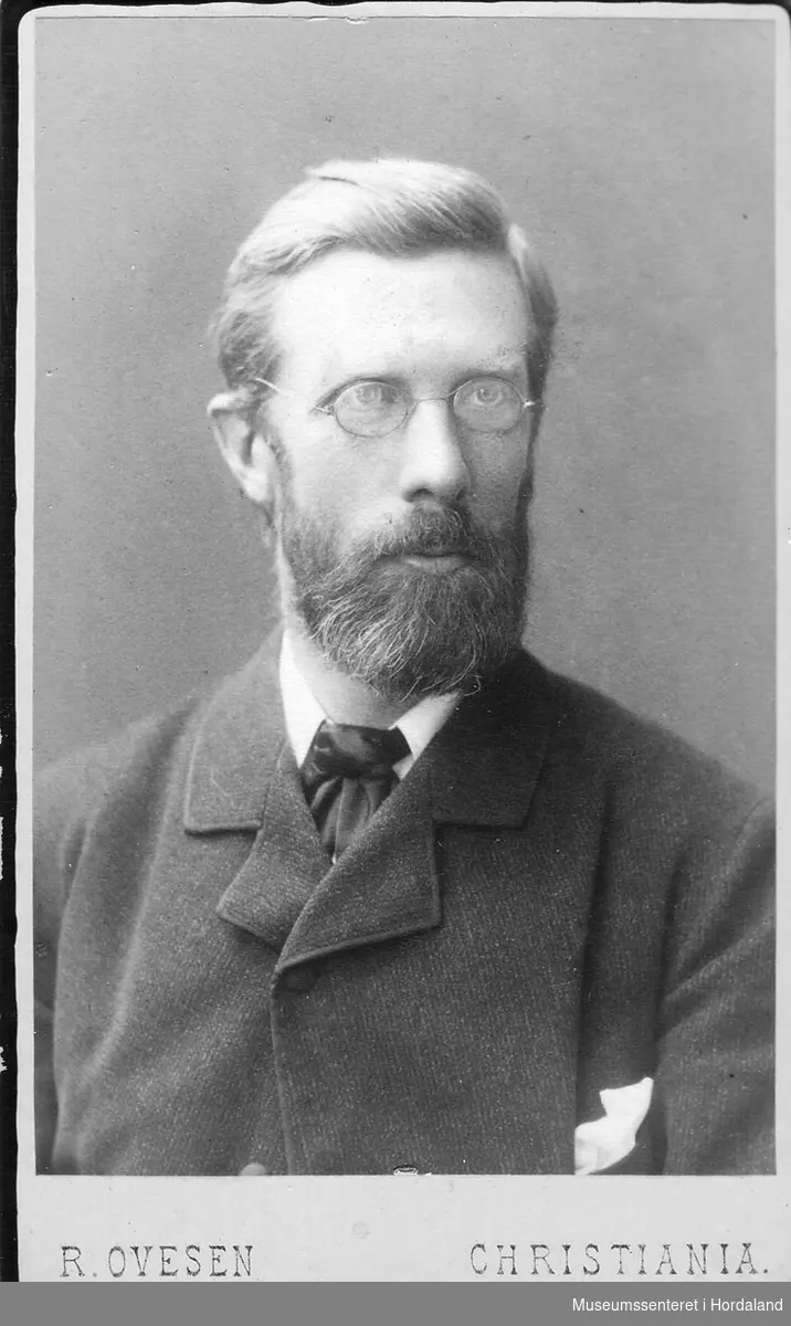 Mann med fullskjegg og runde briller, jakke, skjorte, slips og lommetørkle i brystlomma