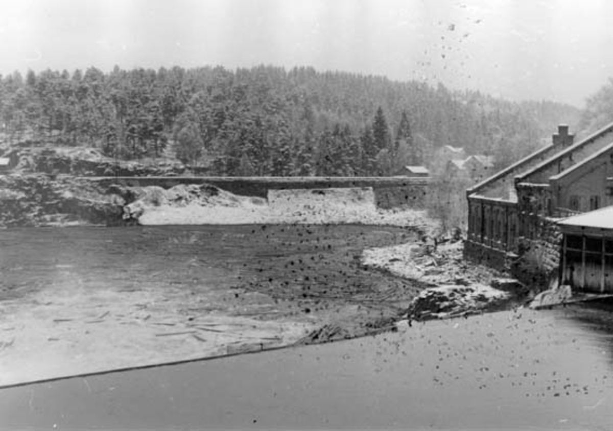 Drammenselvens Papirfabrikker ved Geithusfossen på Modum i Buskerud, fotografert fra øst.  Fotografiet viser den gamle dammen ved fabrikken med restene av det nedbrente nye sliperiet til høyre for elvebredden.  I bakgrunnen ses jernbanetraseen og Horntvetåsen med nysnødekt barskog.  Fotografiet ble tatt i 1952. 