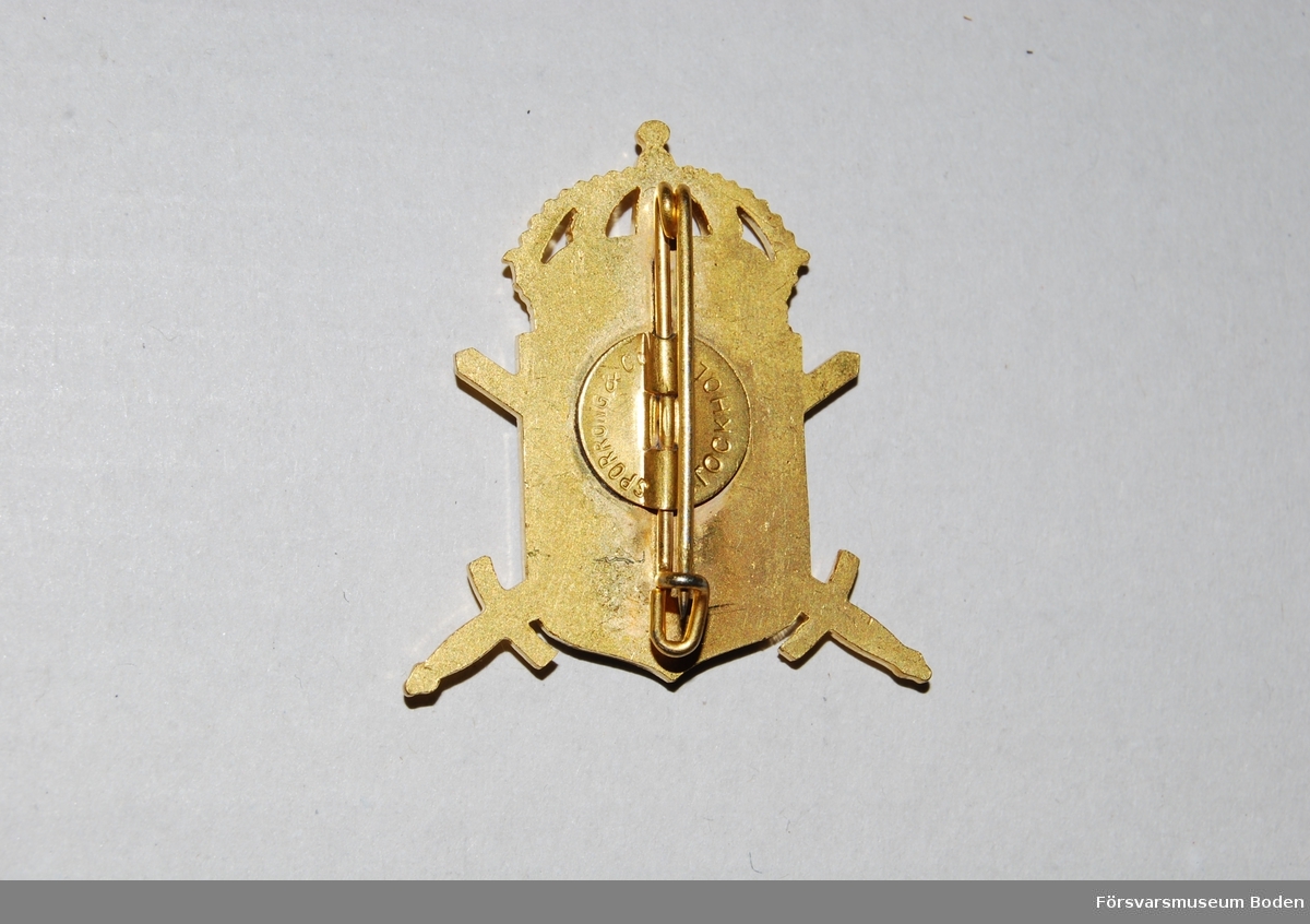 Korslagda svärd i guldfärgad metall med blå sköld vilken är försedd med tre öppna kronor och bokstaven H. Upptill sluten krona. Nål på baksidan.