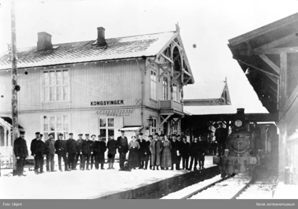 Personale på Kongsvinger stasjon oppstilt foran stasjonsbygningen og med et damplokomotiv, trolig type 4a nr. 20, i spor 1