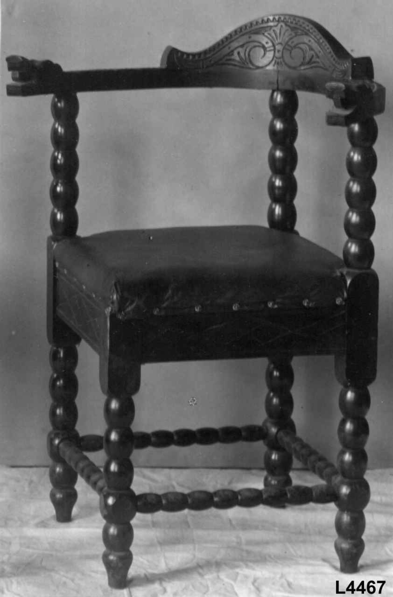 Trestol med diagonalt plassert, firkantet sete med mørkerød voksduk og blomsterformede nagler. Med kuleformer langs bein, sprosser, samt vertikal ryggdel. Øvre del av ryggen er halvsirkelformet og går over til armlenene som har et utskjært dragehode i hver ende.