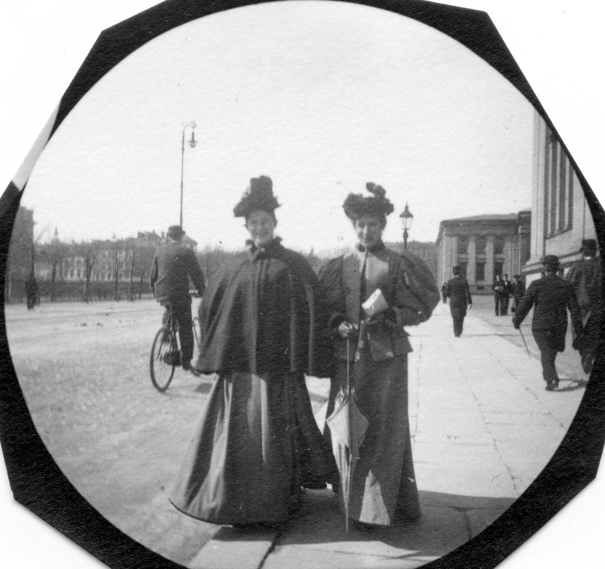To damer spaserer langs Karl Johans gate, Oslo, på fortau utenfor Universitetet, Oslo. Syklist på vei i retning Slottet.