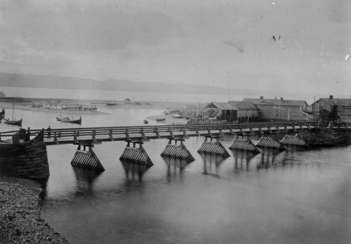 Vestre Jakobselvs, første bro, bygget 1883-1885, Vadsø, Finnmark. Bygninger for enden av broen og båter på elven. Foto fra 1890-årene.